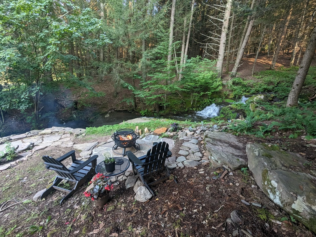 Two black chairs and firepit near a babbling brook with a waterfall at a romantic Catskills getaway cabin.